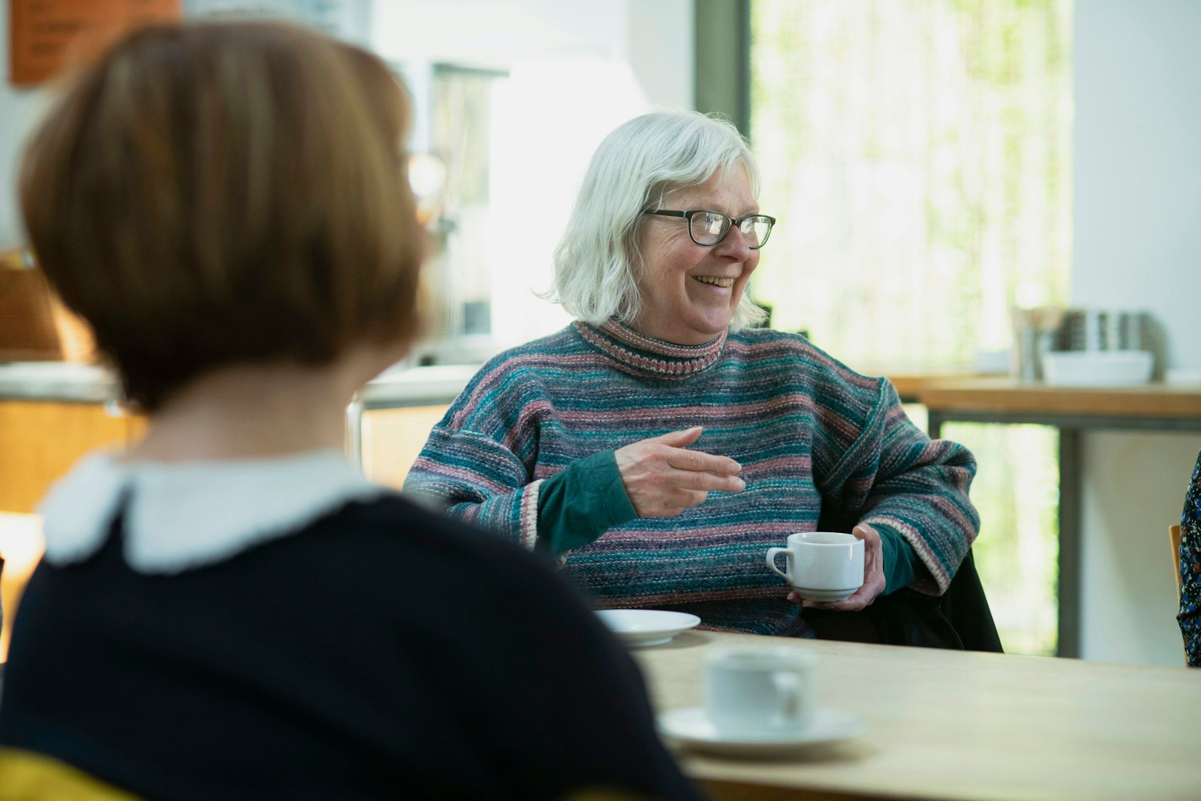 Eine ältere Dame mit Kaffeetasse lacht im Gespräch mit einer Besucherin an einem hellen Küchentisch