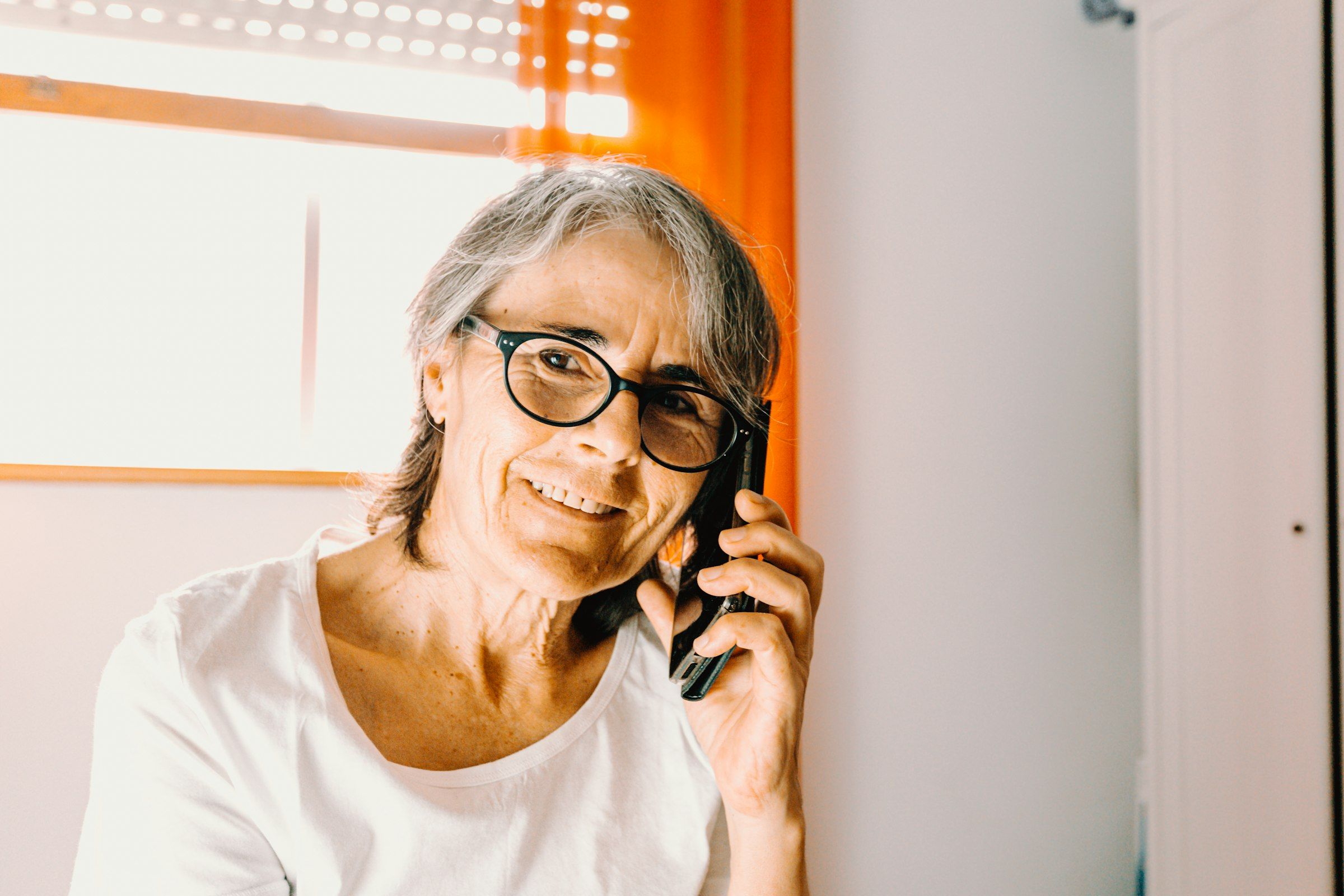 Eine ältere Frau mit Brille telefoniert lächelnd am Fenster